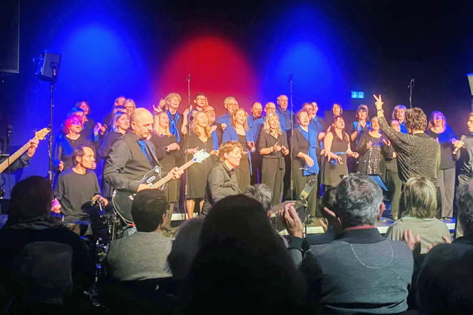 Auftritt eines grossen Gospelchors auf einer Bühne mit blauer und roter Beleuchtung. Im Vordergrund spielt eine Band mit Gitarre, Schlagzeug und Keyboard, während das Publikum applaudiert