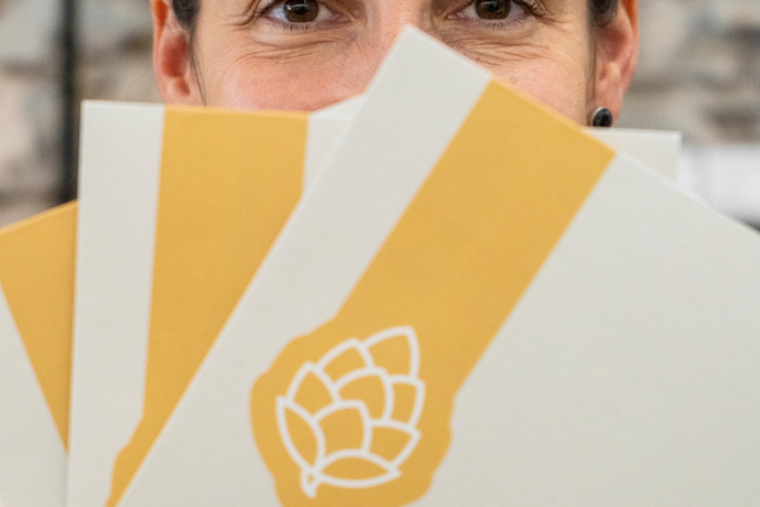 Eine Person hält mehrere gelb-weisse Karten mit Hopfenmotiv fächerförmig vor das Gesicht, sodass nur die Augen sichtbar bleiben.