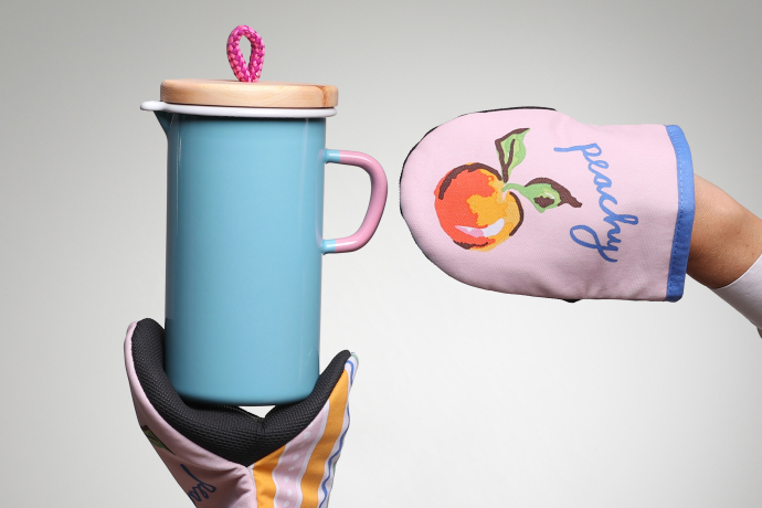 Zwei Hände mit bunten Ofenhandschuhen halten gemeinsam eine hellblaue Kanne mit Holzdeckel; der rechte Handschuh trägt ein Pfirsichmotiv und den Schriftzug ‚peachy‘
