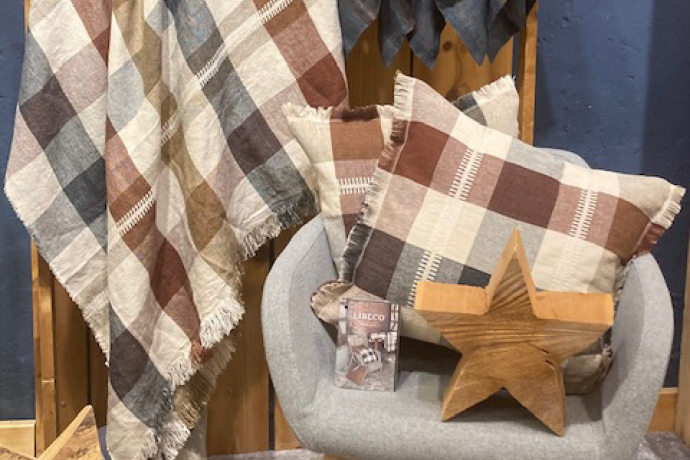 Gemütliche Wohntextilien in warmen Karomustern: Decken und Kissen in Beige-, Braun- und Blautönen sind dekorativ auf einem Sessel und einer Holzrückwand arrangiert, ergänzt durch einen Holzstern.