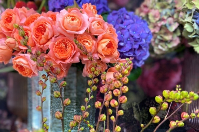 Arrangement aus leuchtend orangefarbenen Rosen, violetten Hortensien und weiteren Blüten in einer strukturierten Vase; im Vordergrund frische Zweige mit grünen und rosafarbenen Knospen.