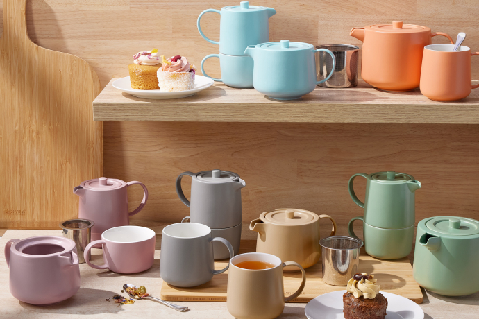 Arrangierte Szene mit farbigen Teekannen und Tassen in Pastelltönen auf Holzregalen und einem Tisch, ergänzt durch kleine Kuchenstücke und Teesiebe