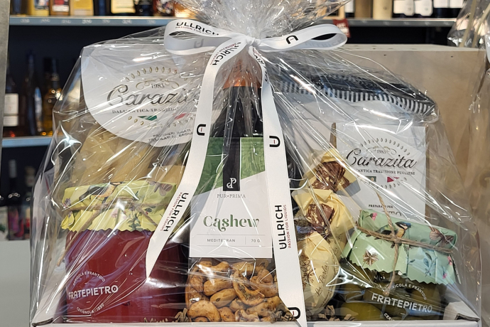 Geschenkkorb mit eingepackten Delikatessen wie Pasta, Olivenöl, Nüssen und Süsswaren, in durchsichtige Folie gewickelt und mit einer weissen Schleife verziert; im Hintergrund Regale mit Spirituosen.