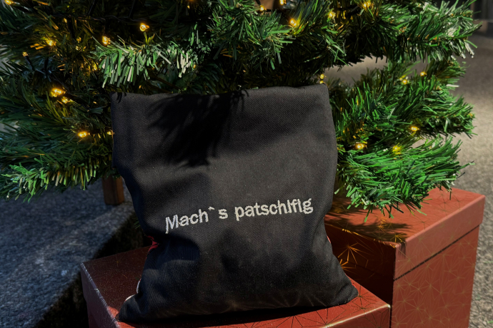 Schwarzes Wärmekissen mit weissem Schriftzug ‚Mach’s patschifig‘, platziert auf einem roten Geschenkpaket unter einem beleuchteten Weihnachtsbaum.
