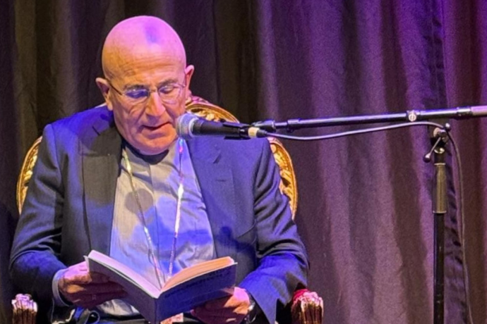 Der Bischof von Chur sitzt auf einem roten, goldverzierten Stuhl und liest bei einer stimmungsvoll beleuchteten Lesung aus einem Buch. Vor ihm stehen Kerzen, Gläser und eine Wasserflasche auf einem Tisch.