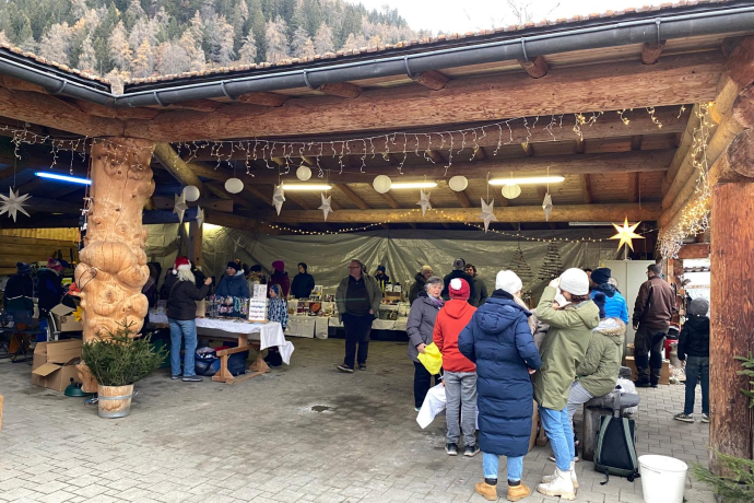 Besucherinnen und Besucher beim Weihnachtsmarkt in Maladers unter einem überdachten Holzpavillon. Verkaufsstände mit regionalen Produkten und Weihnachtsdekorationen, im Hintergrund verschneite Berghänge.