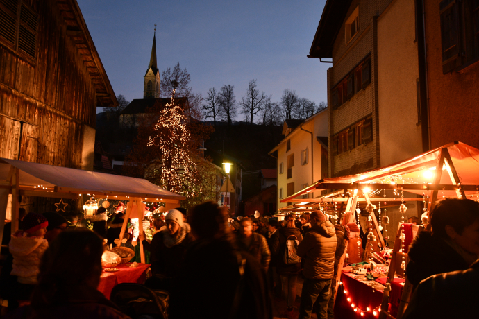 Abendliche Stimmung am Weihnachtsmarkt in Tamins: Zwischen festlich beleuchteten Marktständen schlendern Besucherinnen und Besucher, im Hintergrund ist eine Kirche mit beleuchtetem Turm zu sehen.