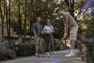 Three people play mini-golf in a sunlit park, surrounded by lush flowers. One is taking a shot while the others watch, creating a relaxed and fun atmosphere