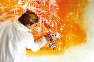 A person in a white coat paints an abstract mural with bold orange and white strokes