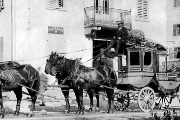 Historisches Schwarz-Weiss-Foto von vier Pferden, die eine Postkutsche ziehen, auf der ein Kutscher sitzt. Drei Männer stehen in der Nähe.