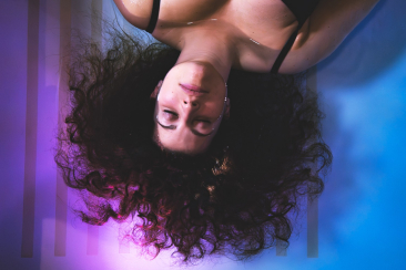 A woman with curly hair lies on her back in a calm, dreamy setting. Her expression is serene, surrounded by soft purple and blue lighting