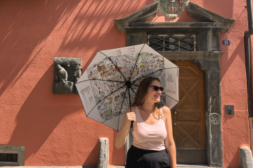 Eine Frau steht vor einer historischen Hausfassade mit roter Wand und altem Holztor. Sie trägt Sonnenbrille, helles Top und dunkle Hose, während sie einen aufgespannten Regenschirm mit Stadtplan-Motiv in der Hand hält und lächelnd zur Seite blickt.