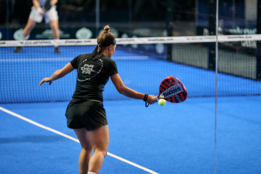 A woman in athletic wear prepares to hit a ball with a paddle on a bright blue padel court. A focused stance and dynamic action convey intensity