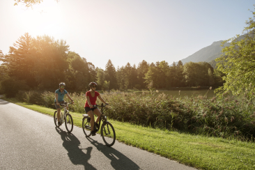 Zwei Radfahrerinnen fahren entspannt auf einem sonnendurchfluteten Weg entlang eines Sees, umgeben von Bäumen und Bergen im Hintergrund. Die Szene vermittelt sommerliche Leichtigkeit und Naturgenuss.