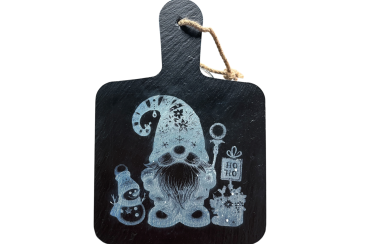 Schieferbrett mit eingraviertem Weihnachtsmotiv: Ein kleiner Wichtel mit Mütze, daneben ein Schneemann, ein Geschenk und ein Schild mit der Aufschrift ‚Ho Ho‘.