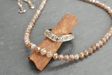 Schmuckarrangement mit naturfarbenen Diamanten: Eine facettierte Diamantkette liegt geschwungen auf dunklem Untergrund, im Vordergrund steckt ein diamantbesetzter Ring auf einer Zimtstange, im Hintergrund hängen passende Ohrringe.