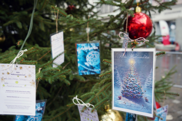 Weihnachtskarten und Wunschzettel hängen an den Zweigen eines festlich geschmückten Tannenbaums im Freien; im Hintergrund sind rote Kugeln und unscharfe Stadtfassaden zu erkennen.