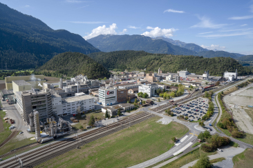 Luftaufnahme des Industrieareals von EMS-Chemie in Domat/Ems mit Produktionsgebäuden, Bahnlinien und Parkplätzen; im Hintergrund bewaldete Hügel und alpine Berge.