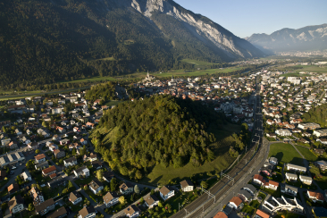 Luftaufnahme von Domat/Ems mit dem bewaldeten Hügel Tuma Casté in der Mitte, umgeben von Wohnhäusern, Bahnlinie und der alpinen Bergkulisse im Hintergrund.
