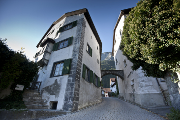 Schmale Gasse im Dorf Felsberg mit historischen Häusern und grünen Fensterläden; ein Steinbogen verbindet zwei Gebäude, im Hintergrund die Calanda-Berge.