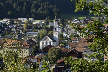 Blick auf das Dorf Rhäzüns mit der Pfarrkirche St. Georg in der Mitte, umgeben von Wohnhäusern und grünen Wäldern am Talrand.