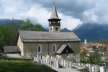 Kirche Sogn Paul in Rhäzüns mit kleinem Friedhof im Vordergrund, dahinter das Dorf und Bergkulisse mit tief hängenden Wolken.