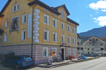 Gelbes historisches Gebäude in Rhäzüns mit Bäckerei, Konditorei und Café im Erdgeschoss, davor Strasse und Blick auf die umliegenden Berge.